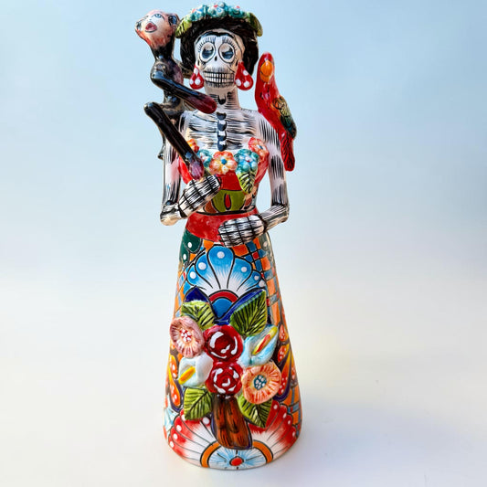 A Talavera Day of the Dead (DĂa de los Muertos) Frida figurine with a monkey on her shoulder and a red parrot on her other shoulder, set against a light blue gradient background.