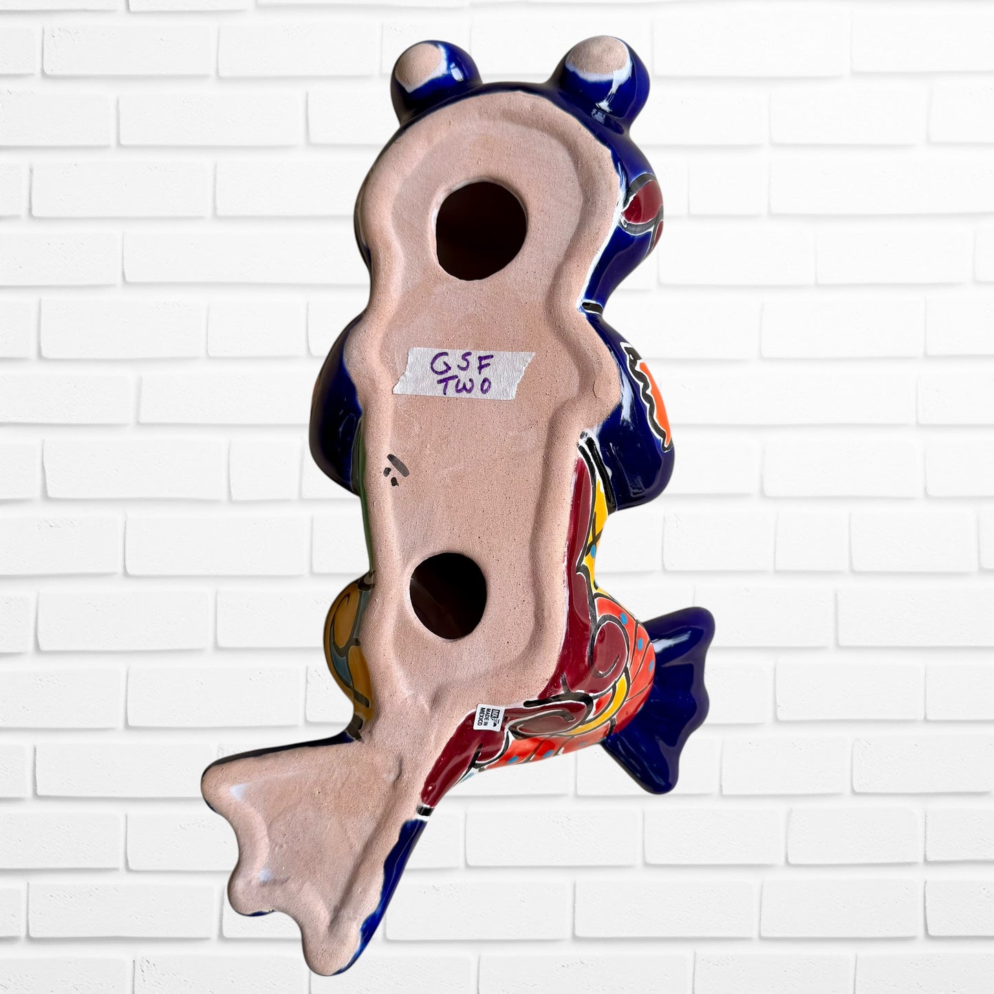 A colorful ceramic frog sculpture sitting cross-legged against a white brick wall background. The frog features a glossy deep blue body with vibrant orange, red, yellow, and light blue painted patterns and details back view with hanging holes.