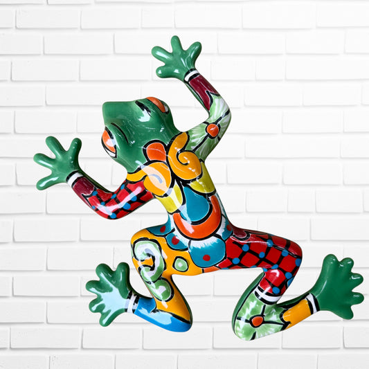 A colorful hand-painted ceramic frog wall sculpture in a dynamic jumping pose, decorated with vibrant green, red, orange, yellow, and blue patterns, set against a white brick wall background.