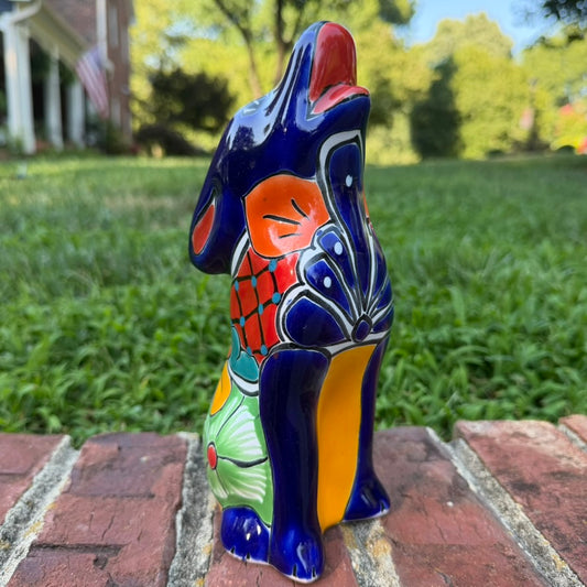 A colorful hand-painted Mexican Talavera Ceramic dog figurine sitting on a brick wall with a green grassy lawn and a house in the background.