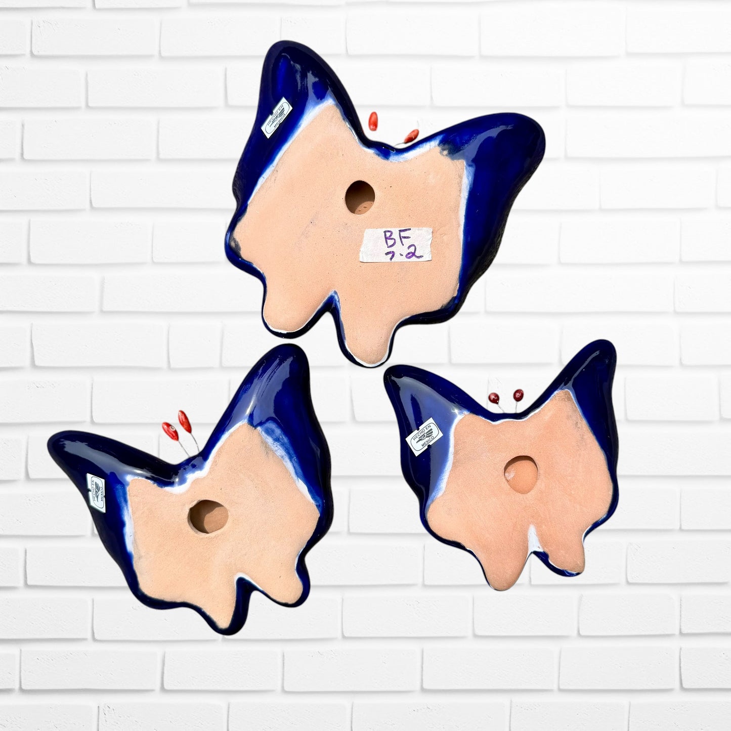 Three colorful ceramic Talavera butterfly wall art pieces with orange, blue, green, yellow, and purple mosaic patterns, displayed against a white brick wall. Back View