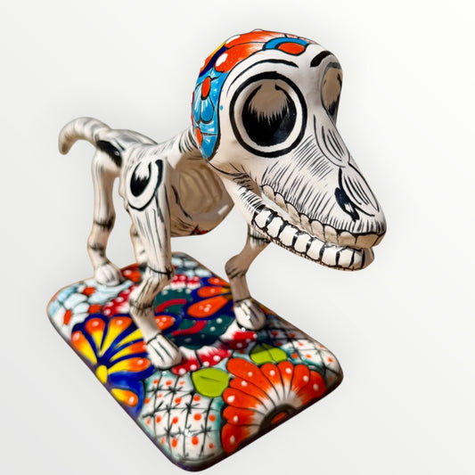 A ceramic Mexican-style skeleton dog figurine standing on a colorful floral base against a plain white background.
