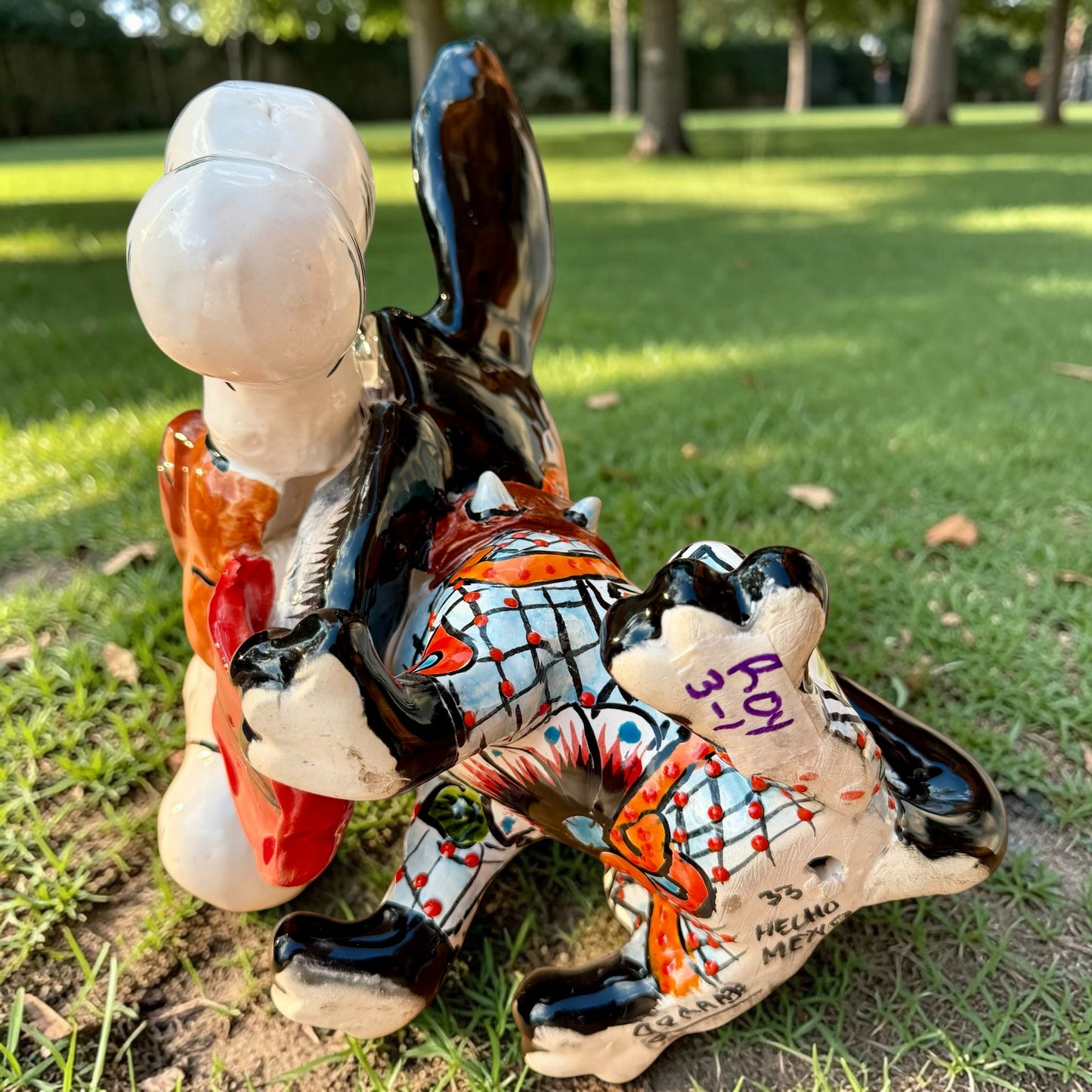 A colorful Talavera dog statue holding a big white bone, photographed outdoors on grass with trees in the background. The dog has a glossy black body, blue eyes, an orange snout, and multicolored patterned ears. Bottom view showing artist signature.