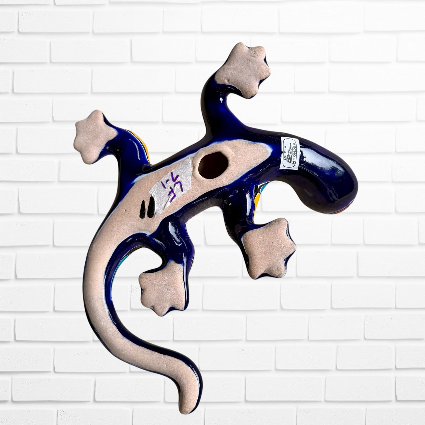 A colorful ceramic wall sculpture of a stylized lizard, decorated with vibrant hand-painted patterns in blue, orange, green, yellow, and red, mounted on a white brick wall bottom view with hanging hole