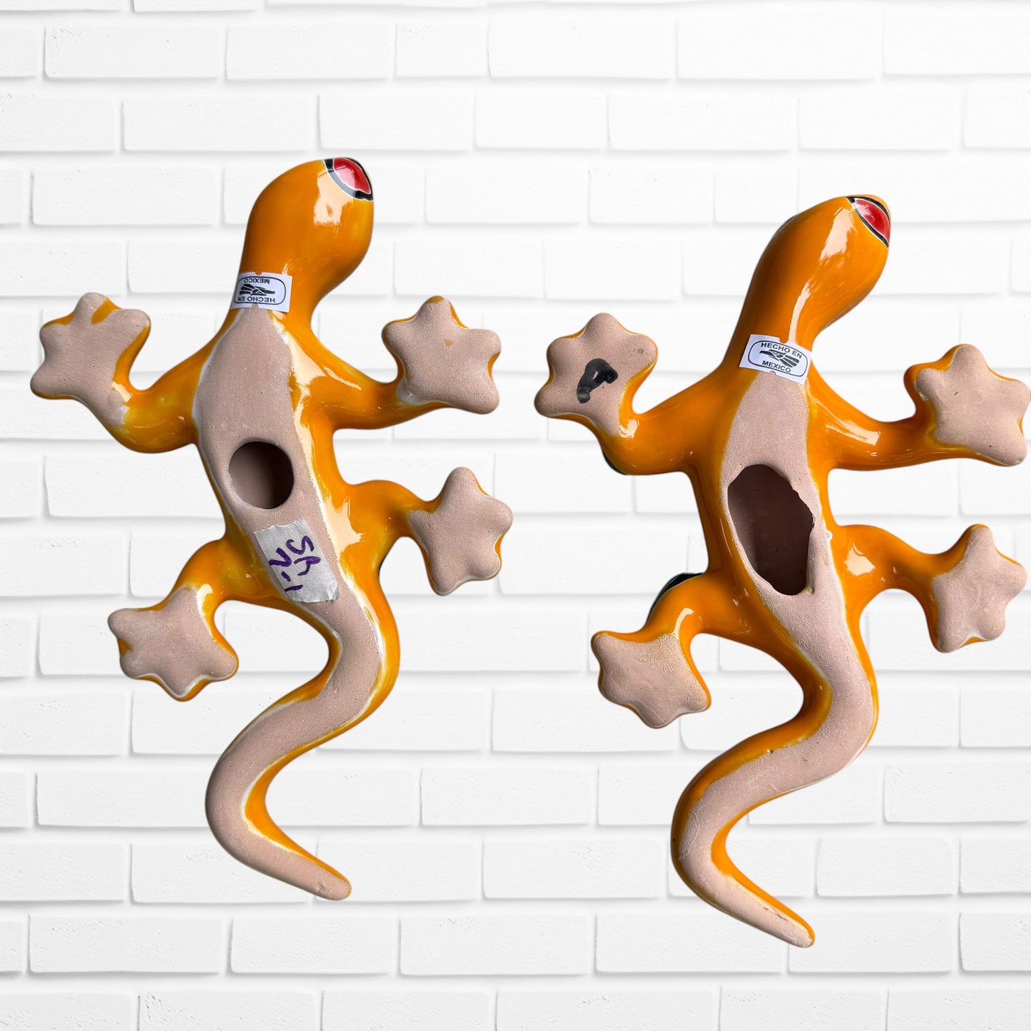 Two colorful hand-painted ceramic gecko wall sculptures in a vibrant Mexican folk art style, displayed side-by-side against a white brick wall back view showing hanging hole.