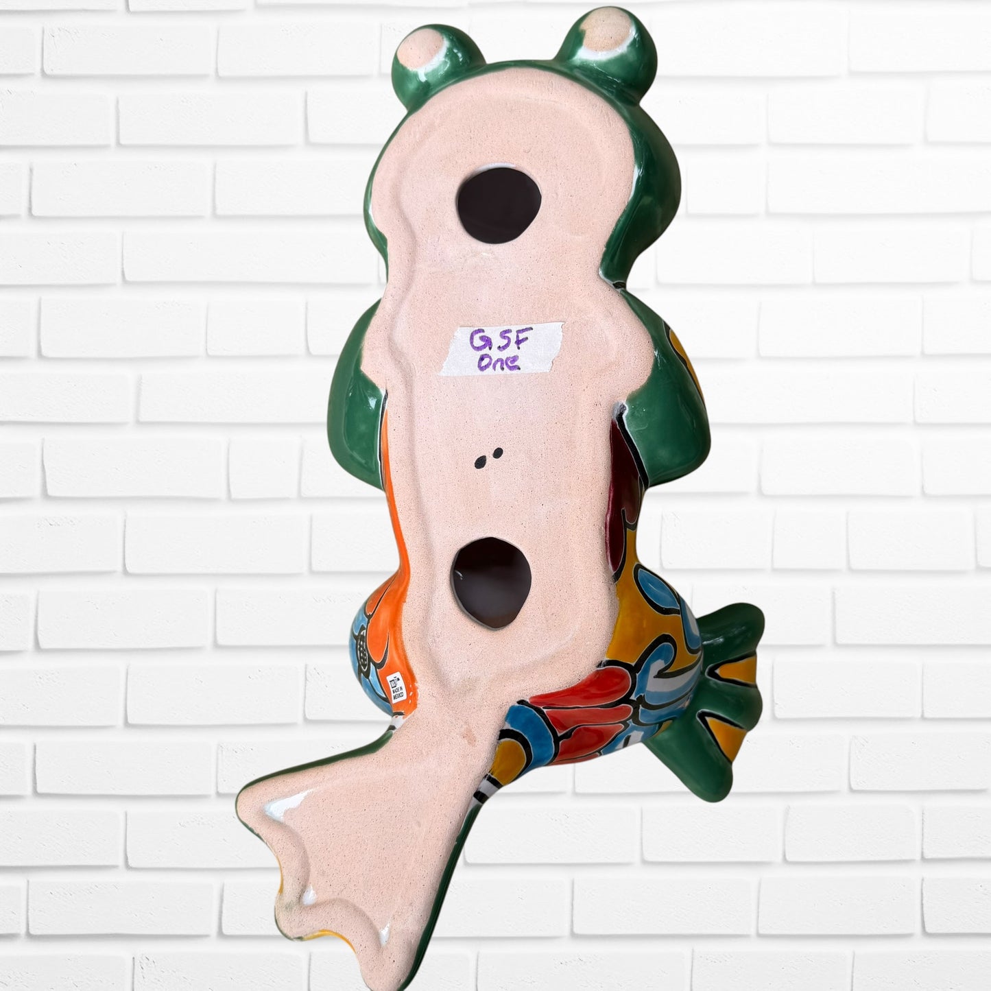 A colorful ceramic frog wall sculpture sitting cross-legged against a white painted brick wall. The frog features a glossy green body with yellow, blue, red, and Back view with hanging holes