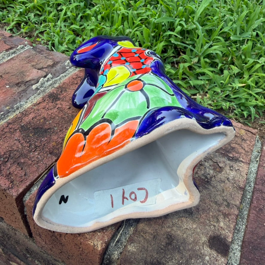 A colorful hand-painted talavera ceramic coyote figurine sitting on a brick surface with a blurred green lawn and trees in the background bottom view