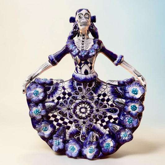 A Day of the Dead style talavera figurine of a skeleton woman in a wide floral skirt, painted in deep blue, black, white, and light blue on a soft beige-to-blue gradient background.