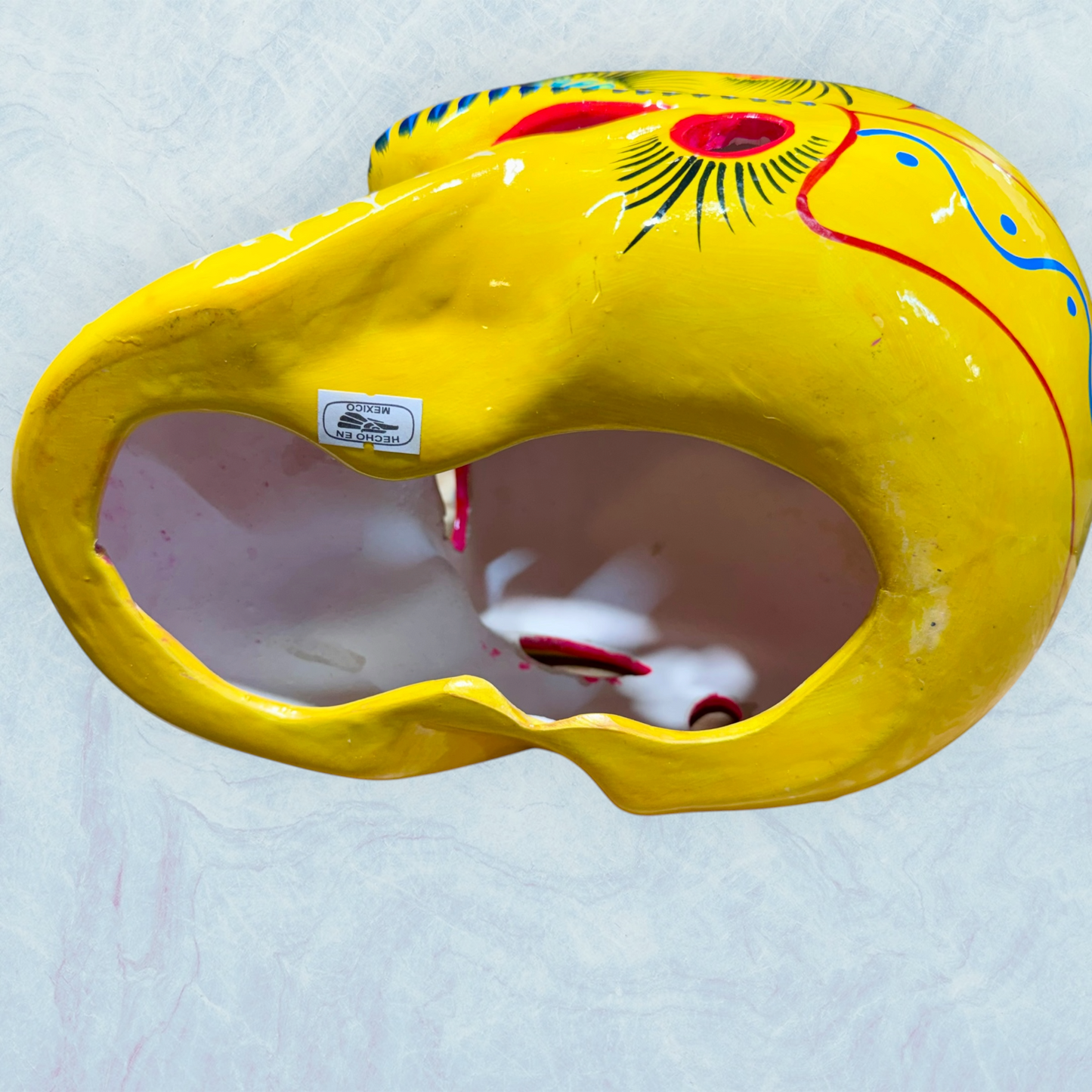 A vibrant yellow sugar skull decorated with colorful floral and geometric patterns in blue, red, black, and white, set against a light blue textured background, bottom view of skull.