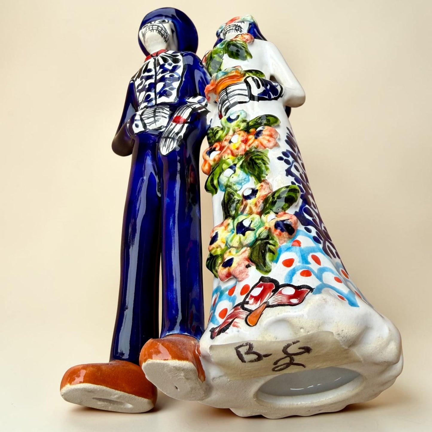 A hand-painted Talavera Day of the Dead bride and groom figurine. The groom is dressed in a blue suit with white patterns, a black bow tie, and an orange-red bow tie accent, wearing a blue hat. Bottom view.