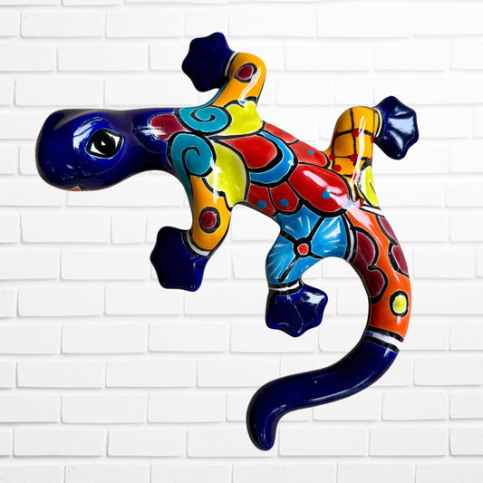A colorful ceramic wall sculpture of a stylized lizard or salamander, decorated with vibrant hand-painted patterns in blue, red, yellow, and orange, mounted on a white brick wall.