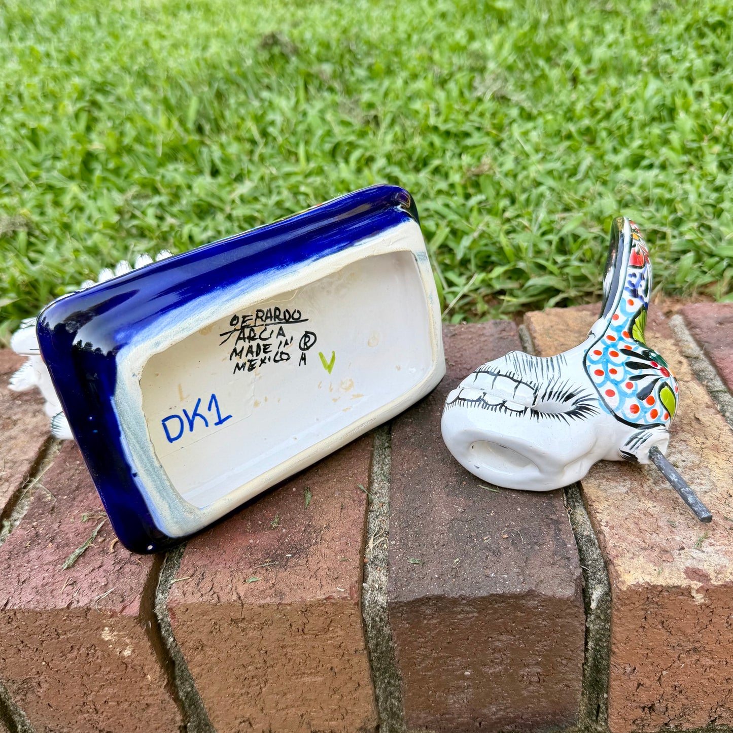 A hand-painted Mexican Talavera-style donkey figurine with a skeletal design, standing on a colorful floral base outdoors on a brick path with a green lawn in the background. This is the bottom view with artist signature.