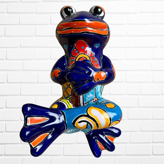 A colorful ceramic frog sculpture sitting cross-legged against a white brick wall background. The frog features a glossy deep blue body with vibrant orange, red, yellow, and light blue painted patterns and details.