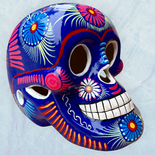 A glossy blue Day of the Dead sugar skull XGrande decorated with vibrant pink, blue, white, and orange floral and geometric patterns, shown against a light blue textured background.
