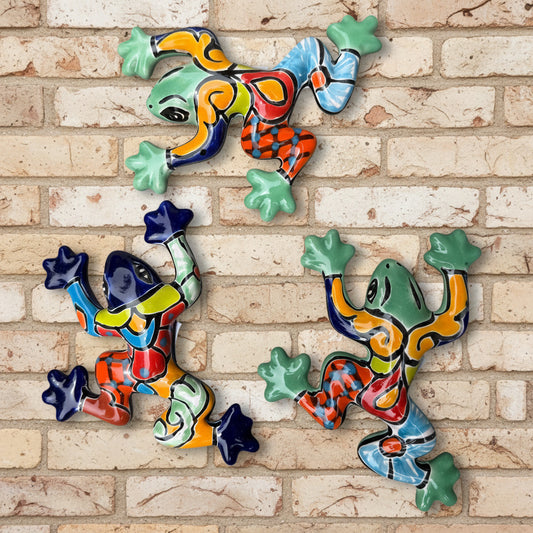 Three colorful ceramic wall-mounted frog sculptures in a vibrant multicolor design, displayed against a white brick wall background.