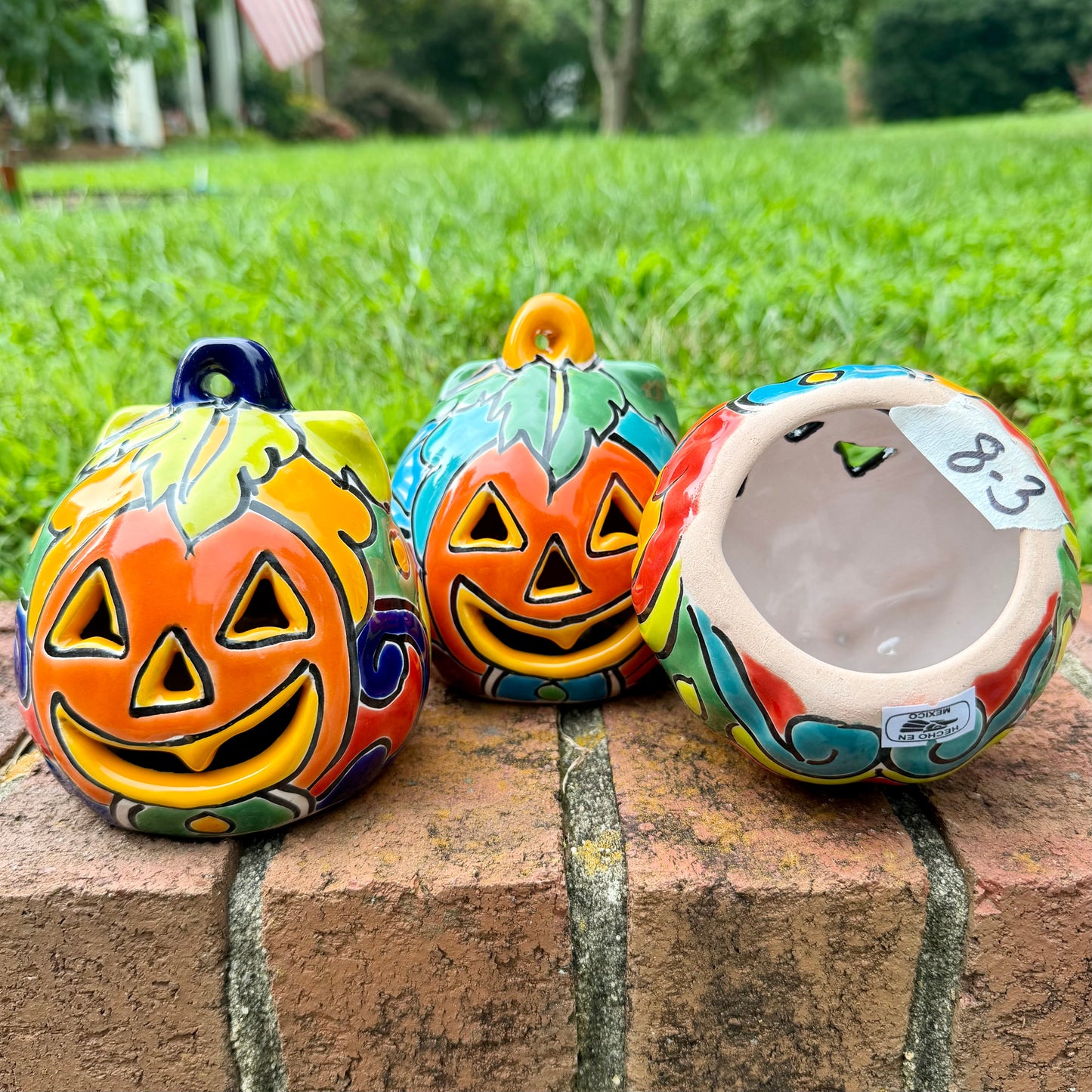 Three hand-painted Talavera ceramic jack-o'-lantern pumpkins with orange faces and yellow, blue, and red green designs, sitting on a brick path in a grassy backyard, bottom view.