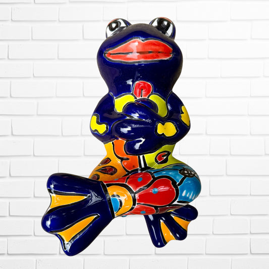 A colorful ceramic frog figurine sitting with arms crossed, decorated with vibrant hand-painted patterns in blue, yellow, red, orange, and light blue, set against a white brick wall background.