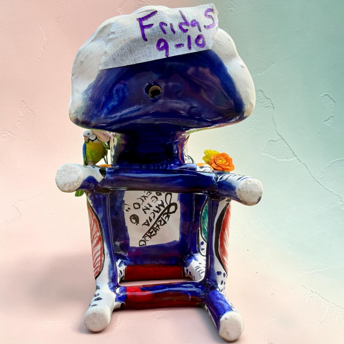 Frida Kahlo Talavera day of the dead figurine sitting in a chair holding a monkey with a parrot on her shoulder. She is painted and bright colors and signed by the artist. Photo of the signature on the bottom of the chair.