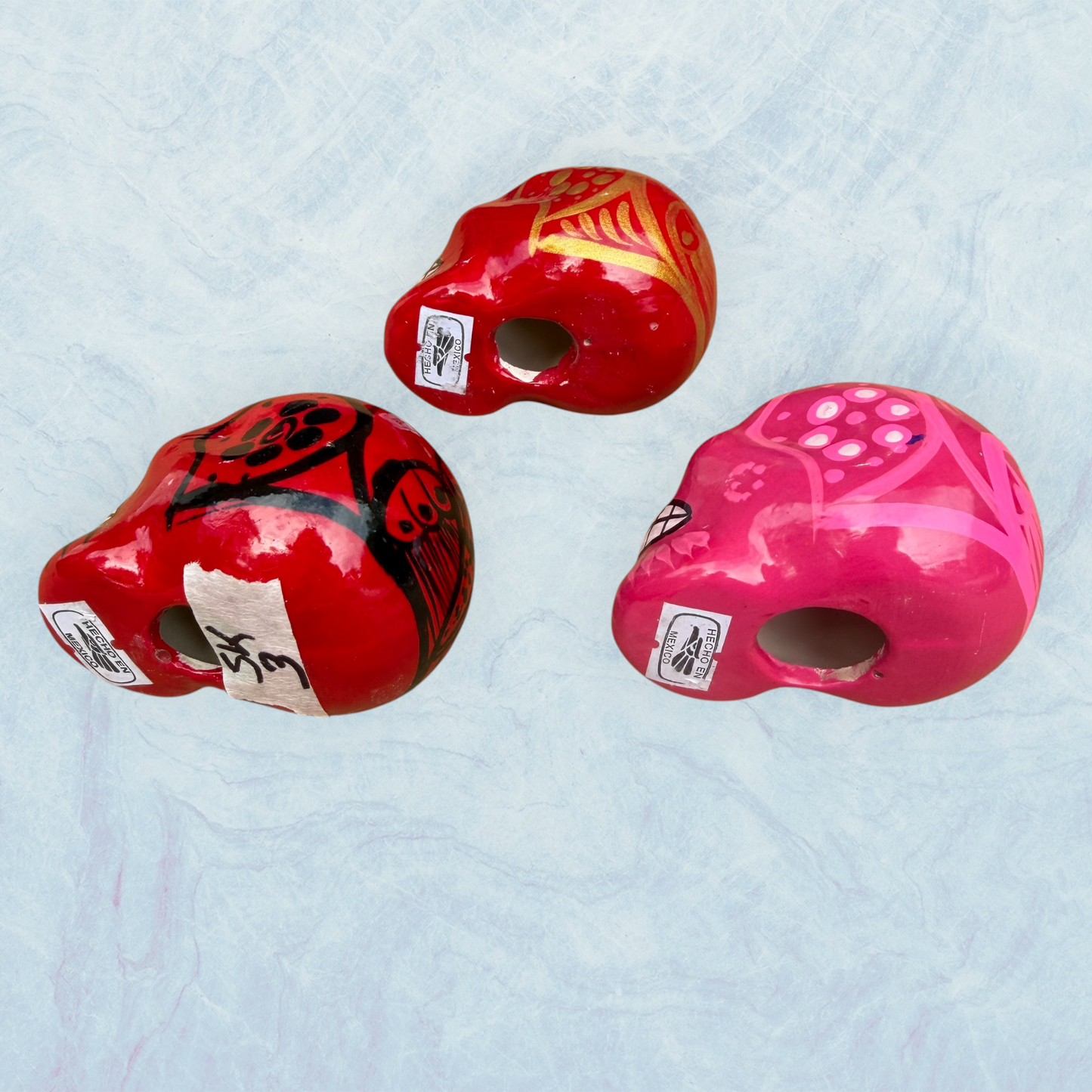 Three hand-painted Mexican sugar skulls (calaveras) in red, orange, and pink, decorated with intricate black and white patterns, set against a light blue textured background, bottom view.