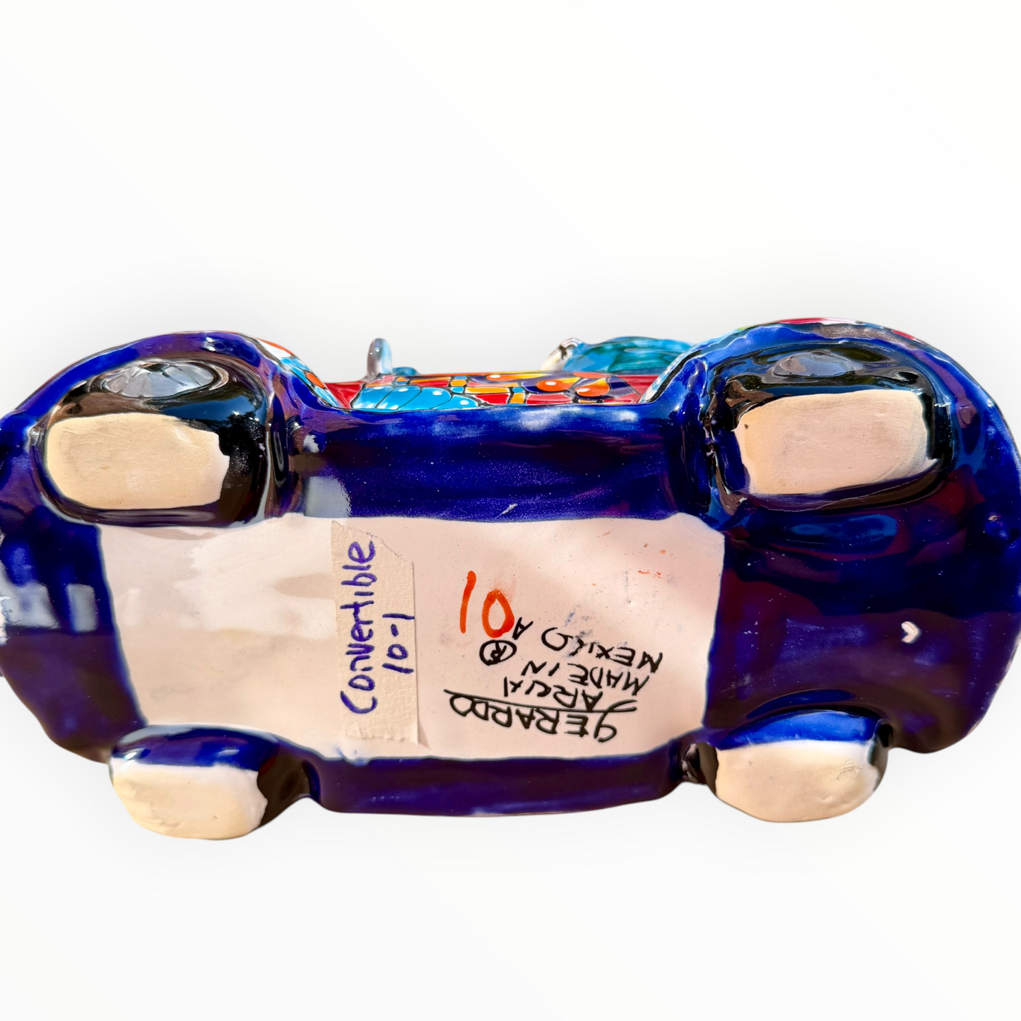 A colorful hand-painted ceramic Mexican-style convertible car figurine with Day of the Dead skeleton characters inside, shown on a neutral beige background. Bottom view with signature.