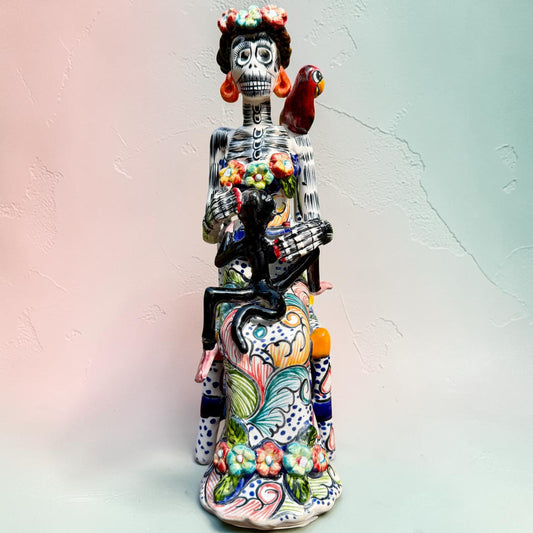 Frida Kahlo Talavera day of the dead figurine sitting in a chair holding a monkey with a parrot on her shoulder. She is painted bright colors and signed by the artist.
