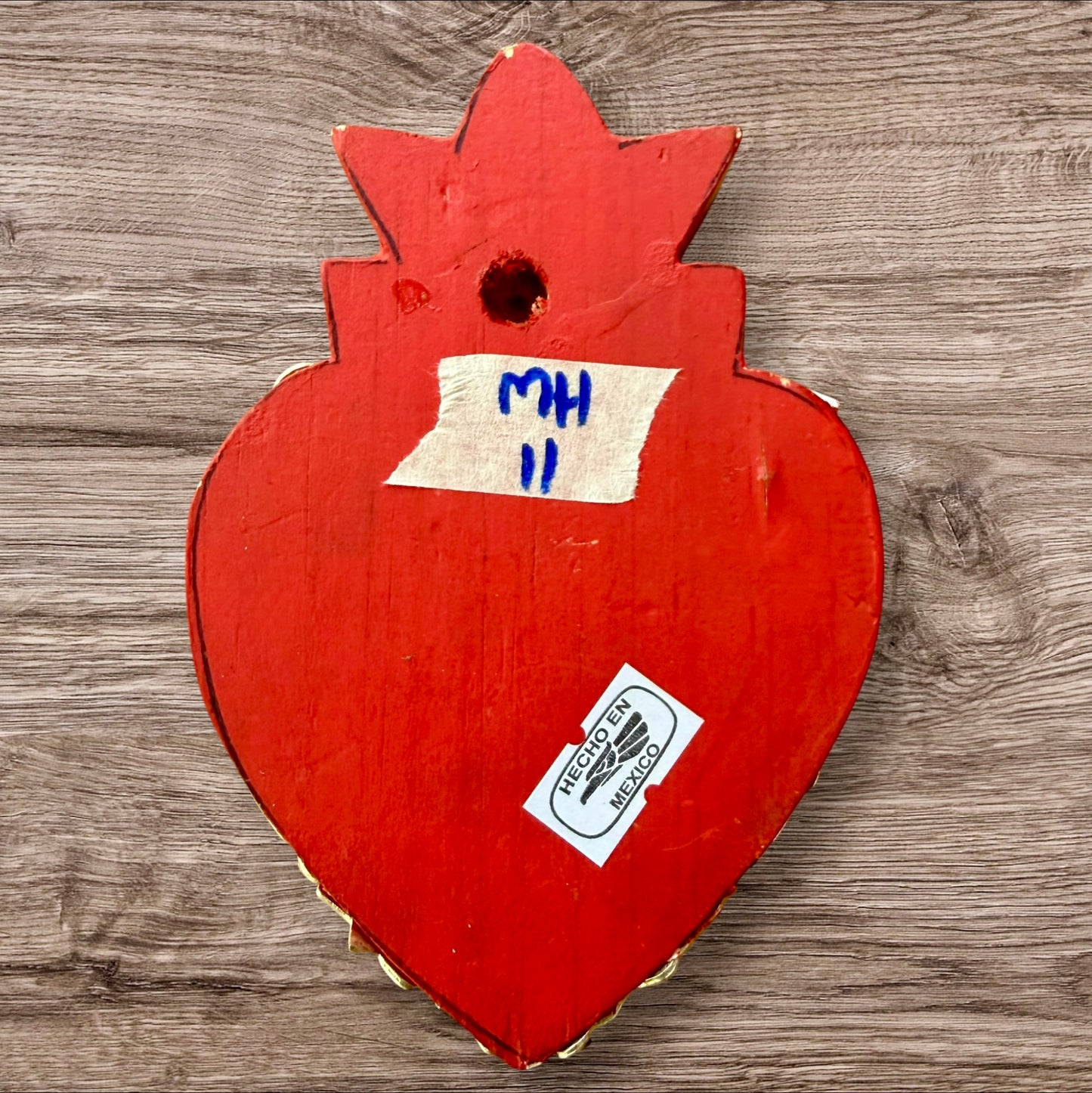 A Milagros heart featuring a gold textured top and a black base covered in raised silver-toned charms displayed on a wooden wall, back view.