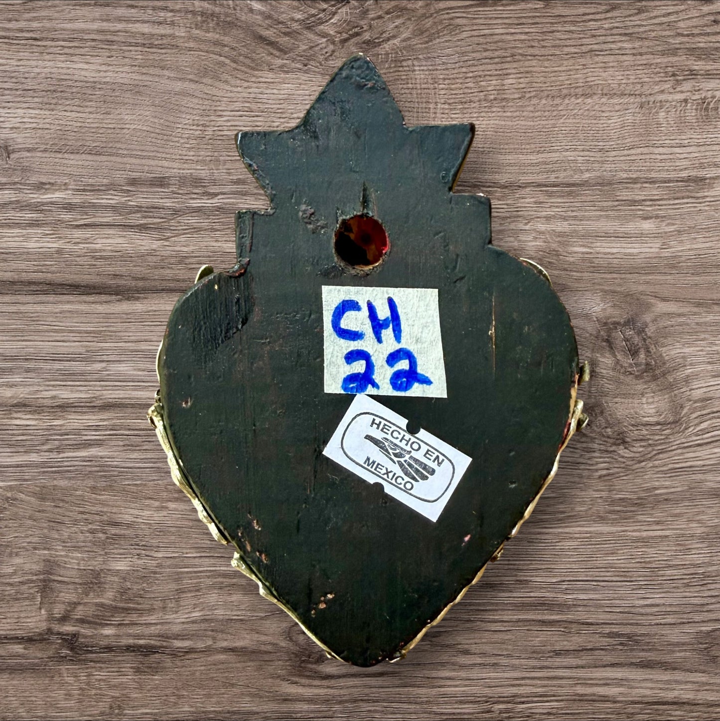 A decorative Milagros Sacred Heart featuring a black and silver painted heart adorned with charms, set against a rustic wood background, this is the back view.