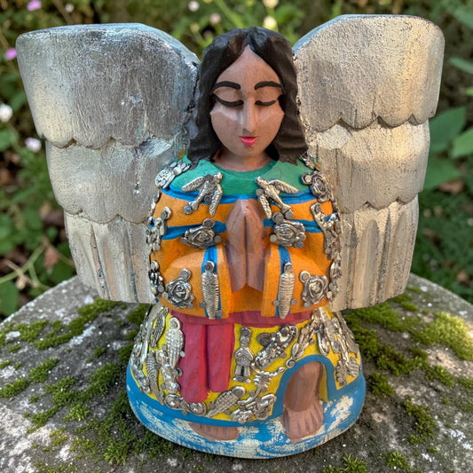 Milagros wooden angel figurine with silver wings and metallic charms, displayed outdoors on a mossy stone with green foliage in the background. The angel is in preyer.