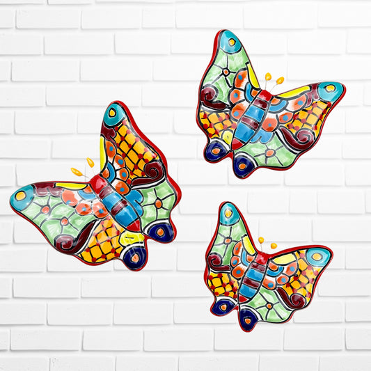Three colorful ceramic Talavera butterfly wall art pieces with orange, blue, green, yellow, and purple mosaic patterns, displayed against a white brick wall.