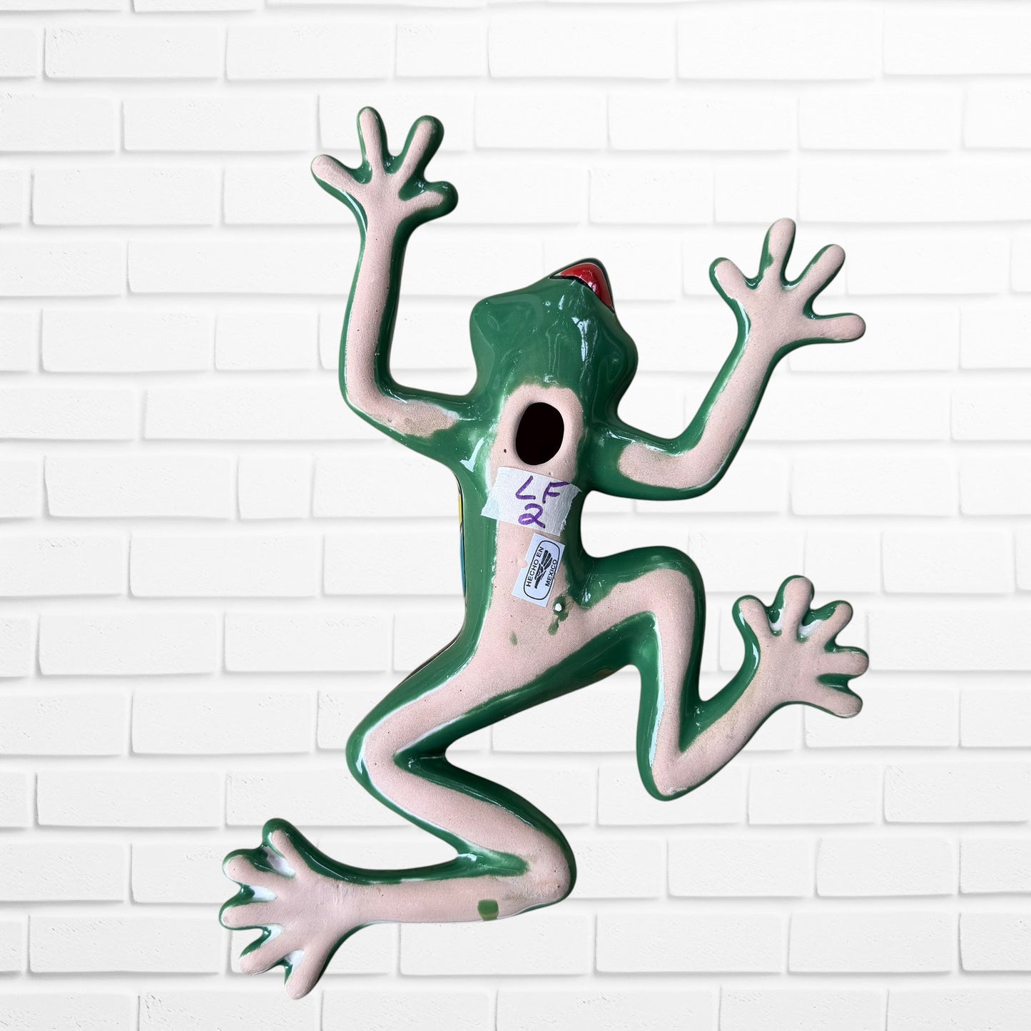 A colorful hand-painted ceramic frog wall sculpture in a dynamic jumping pose, decorated with vibrant green, red, orange, yellow, and blue patterns, set against a white brick wall background back.