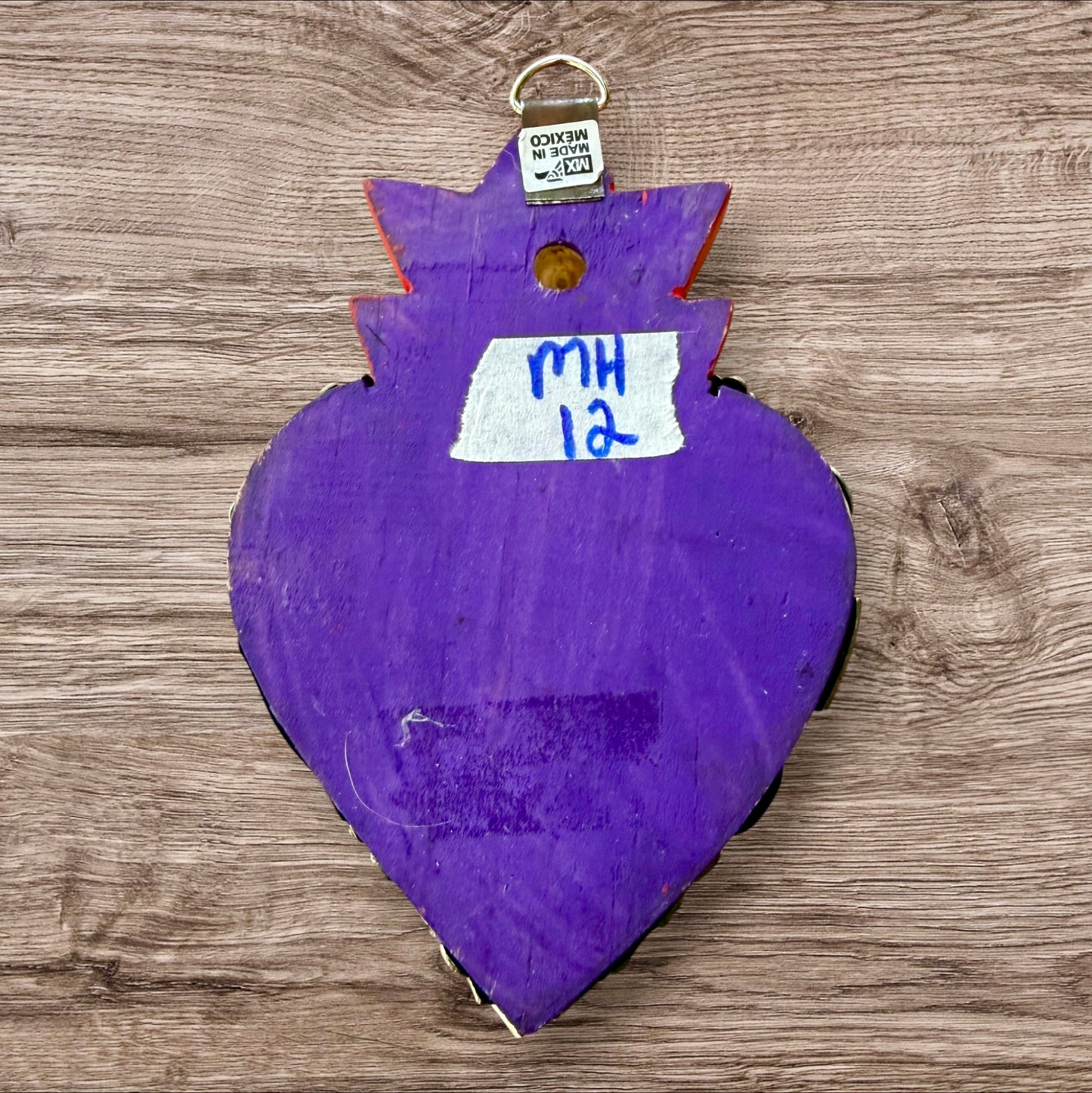 A Milagros heart featuring a gold textured top and a purple base covered in raised silver-toned charms displayed on a wooden wall.