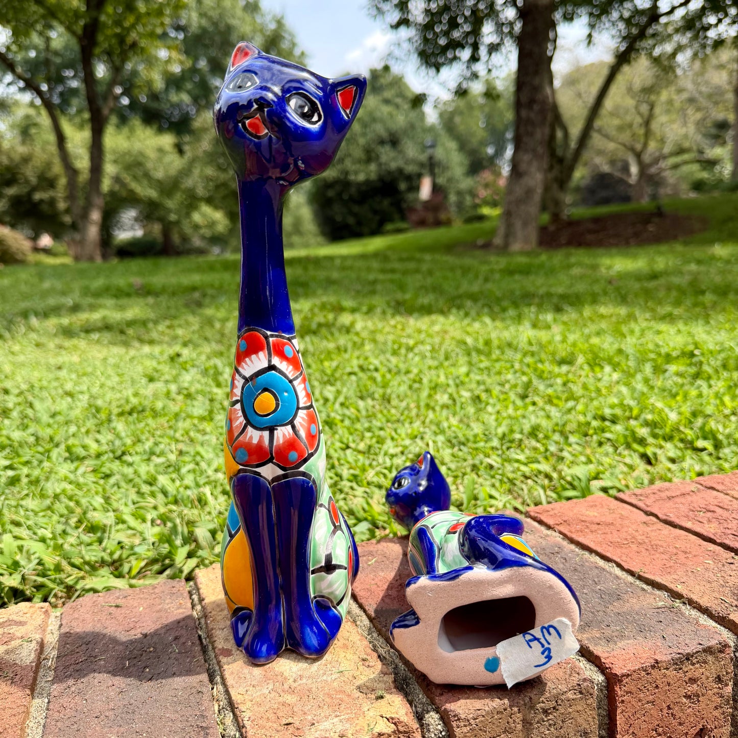 Two colorful hand-painted ceramic cat figurines displayed outdoors on a brick path with a green grassy lawn and trees in the background. One figurine is standing upright and is deep cobalt blue with red, orange, yellow, and white painted patterns on its body.