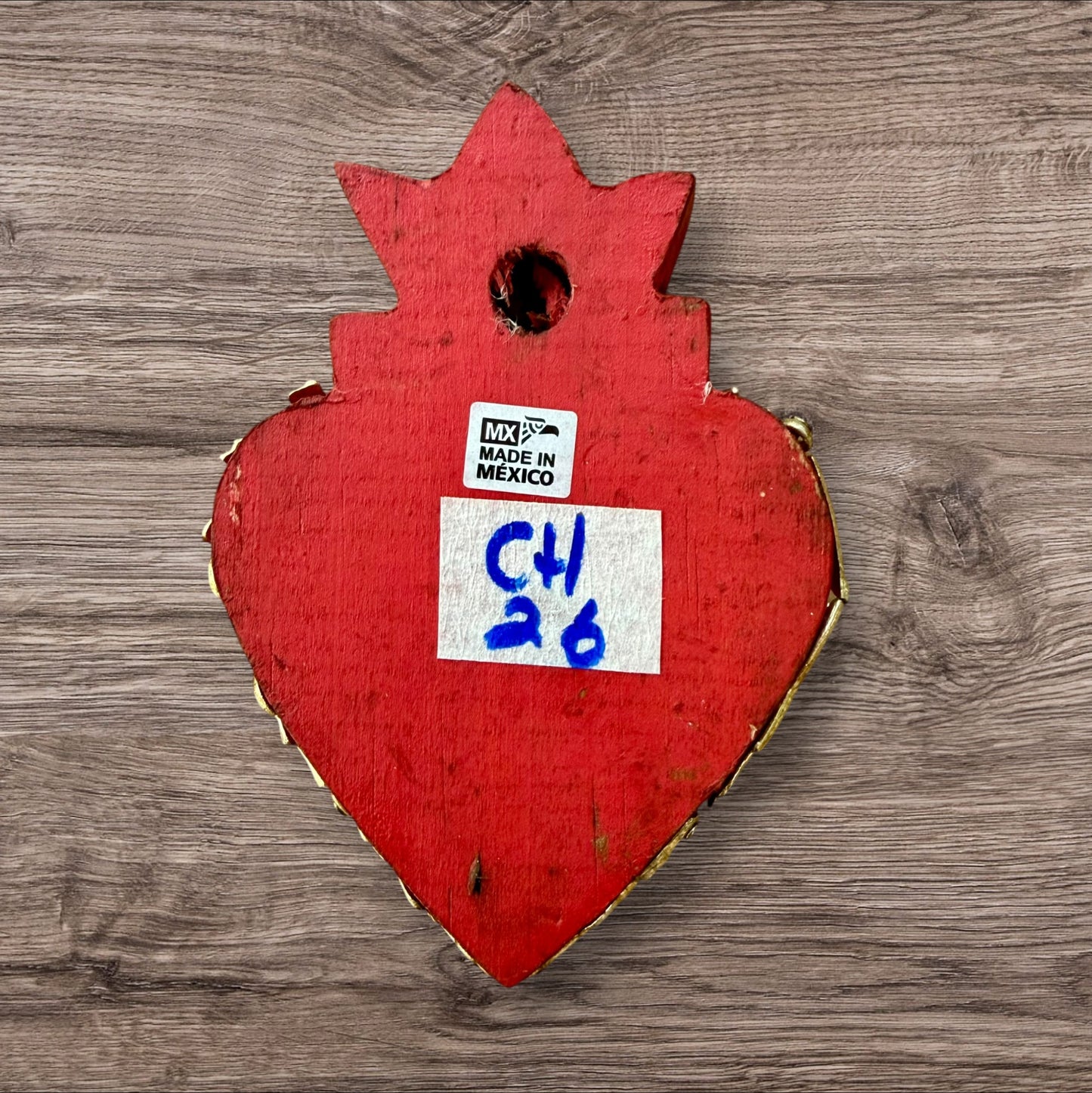 A decorative Milagros Sacred Heart featuring a red and gold painted heart adorned with charms, set against a rustic wood background, this is the back view.