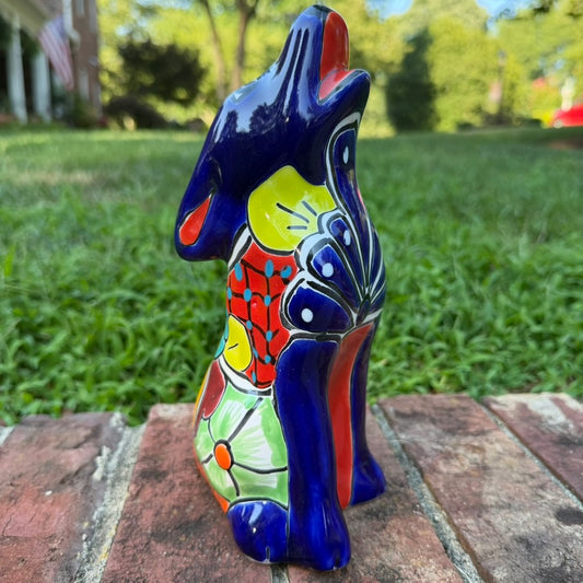 A colorful hand-painted talavera ceramic coyote figurine sitting on a brick surface with a blurred green lawn and trees in the background.