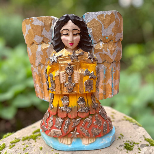 A handcrafted wooden Milagros angel figurine with large wings, decorated with metallic silver charms and painted in yellow, and orange, standing on a mossy stone with a blurred green garden background.
