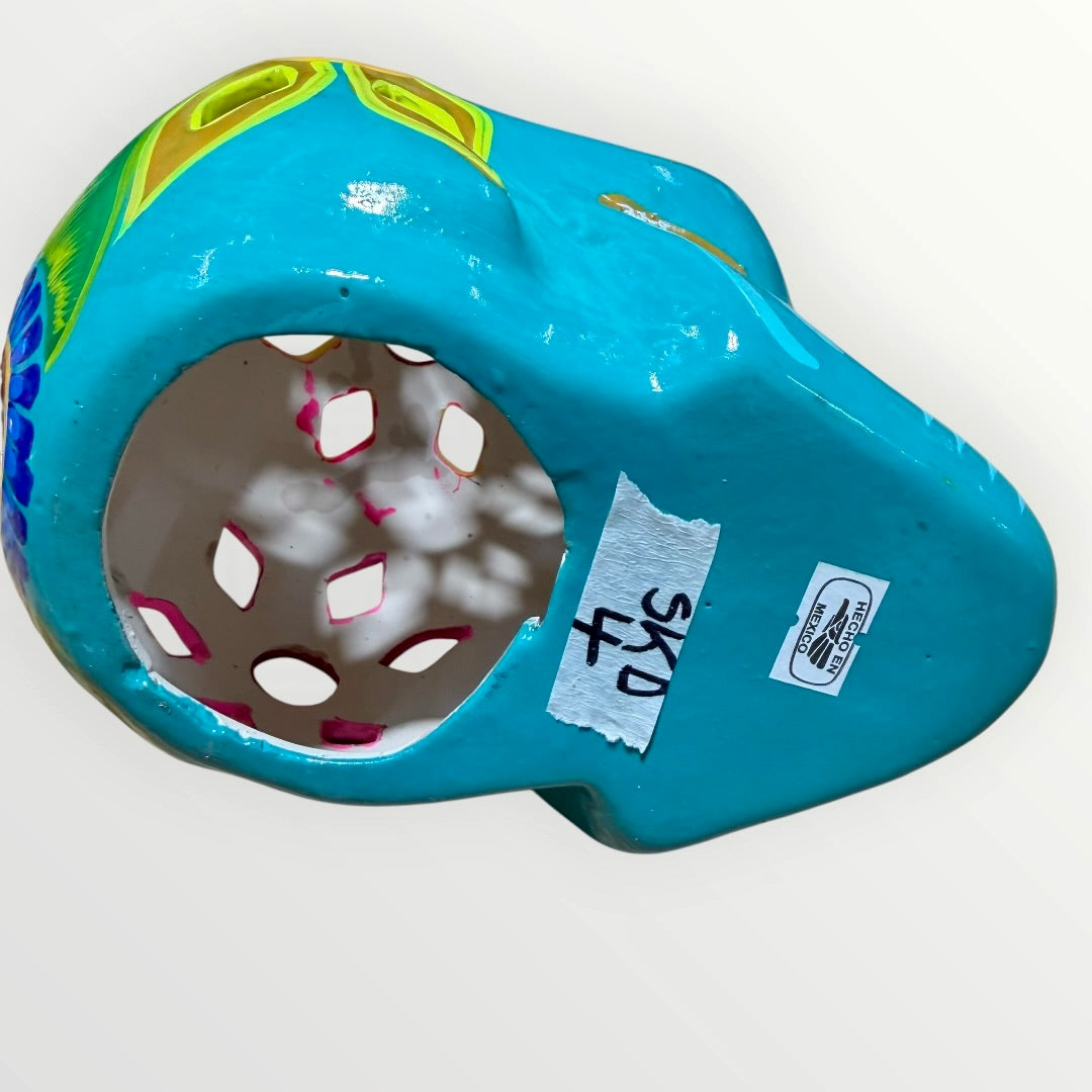A decorative sugar skull painted in bright turquoise blue with colorful floral and geometric patterns in pink, yellow, green, purple, and white, shown against a plain light background bottom view.