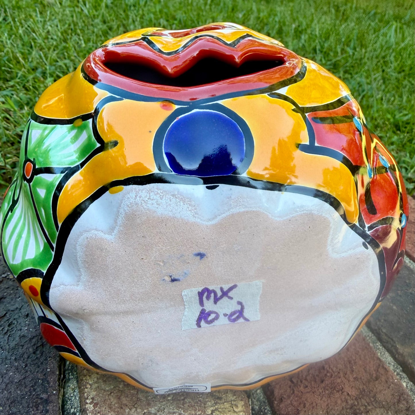 A hand-painted talavera jack-o'-lantern pumpkin garden ornament sitting outdoors on a brick path with green grass and trees in the background. The pumpkin features a bright yellow face with red and black painted facial features, a blue stem, and multicolored painted accents on top. This is the bottom view.