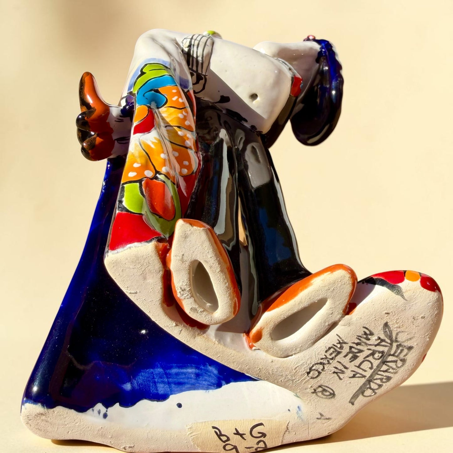 A colorful Day of the Dead figurine of a skeleton bride and groom embracing, with the groom holding the bride. The figurine features vibrant painted details including a orange and white patterned dress, a multicolored hat, and dark hair, set against a plain light background. Bottom view showing signature.