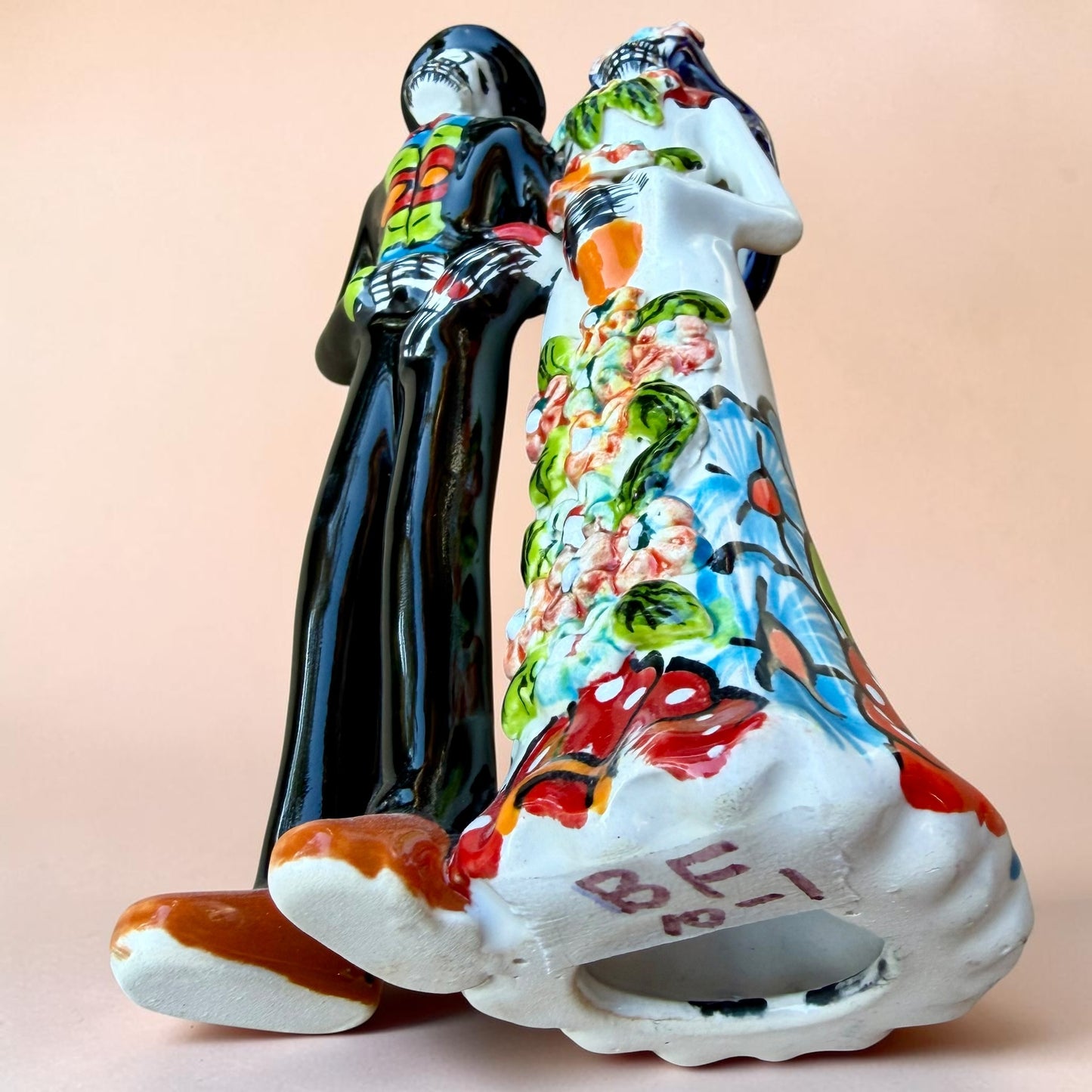 A talavera Day of the Dead wedding couple figurine featuring a groom skeleton in a black suit with colorful floral patterns and a bride skeleton in a white wedding gown with floral details, set against a solid pale peach background. Bottom View.