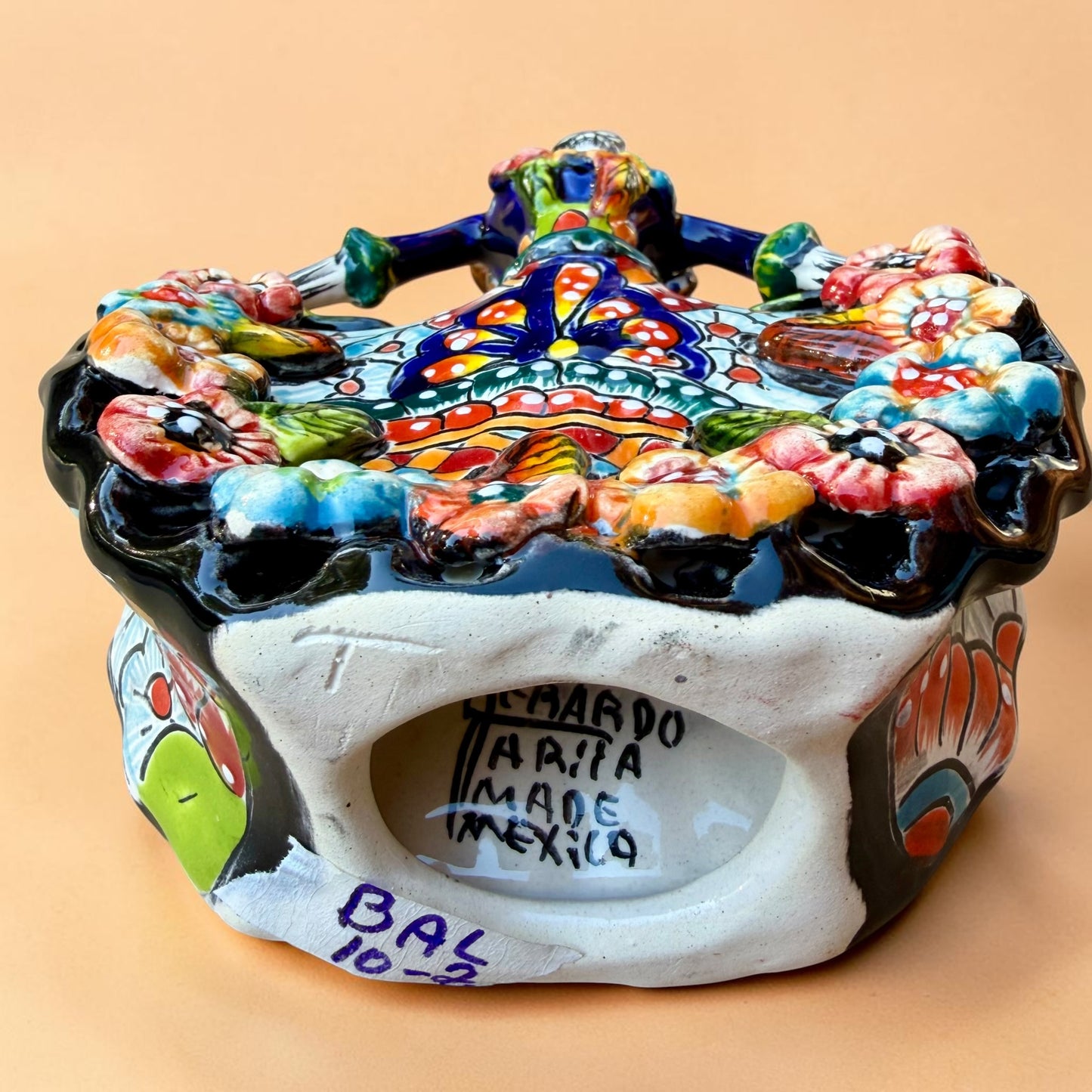 A colorful hand-painted Day of the Dead (Día de los Muertos) ceramic figurine of a ballerina, decorated with vibrant floral patterns and a white skull head with braided hair, set against a solid peach background. Bottom view showing signature.