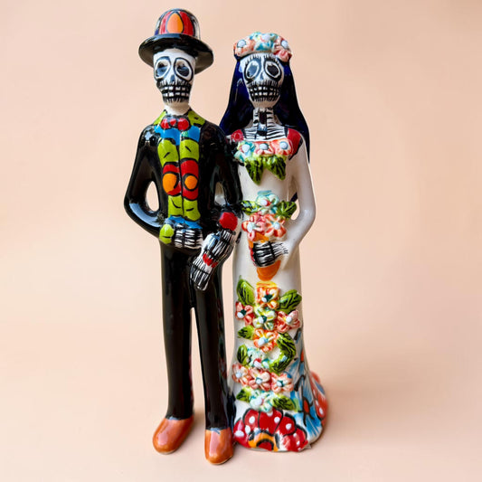 A talavera Day of the Dead wedding couple figurine featuring a groom skeleton in a black suit with colorful floral patterns and a bride skeleton in a white wedding gown with floral details, set against a solid pale peach background.