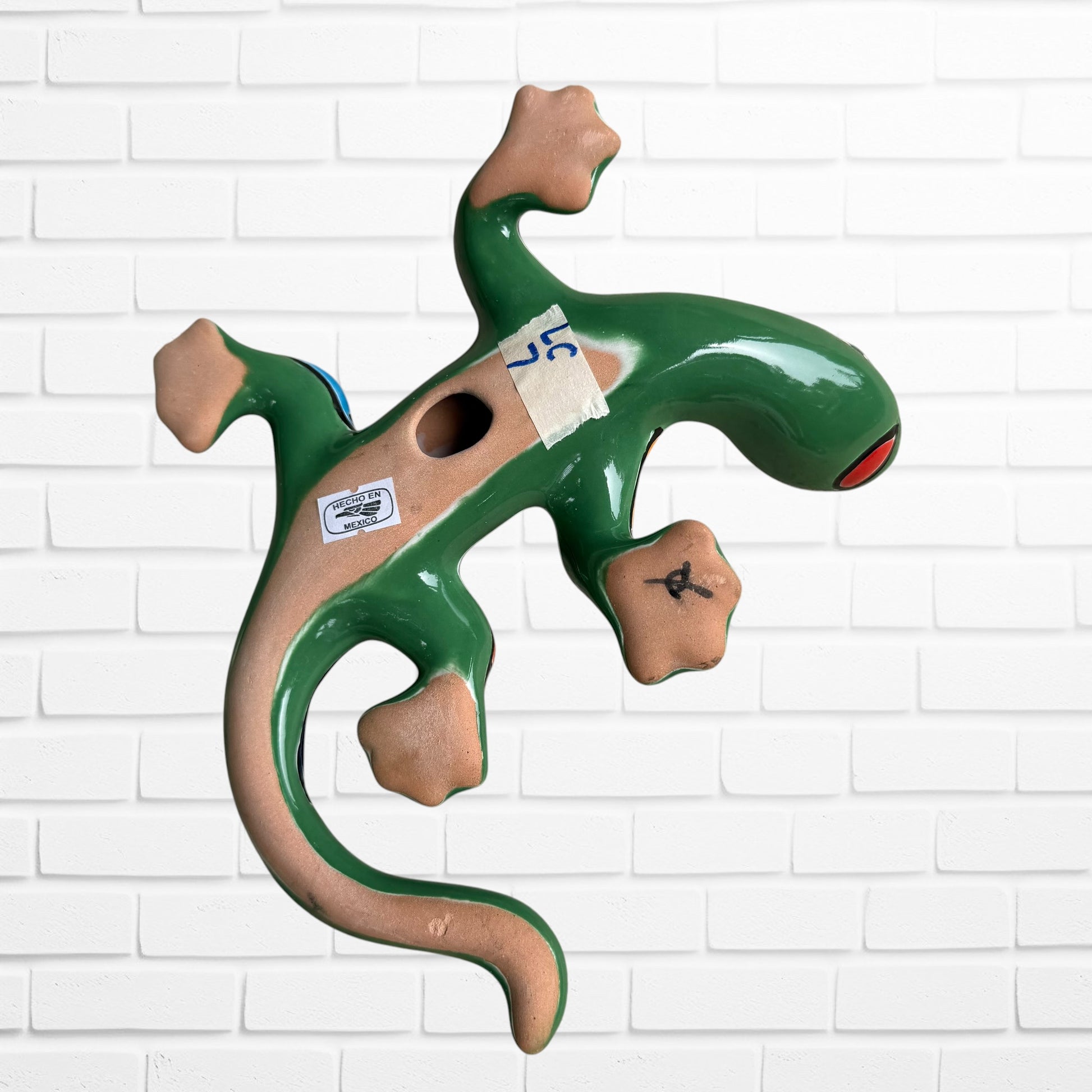 A colorful hand-painted ceramic lizard wall sculpture mounted on a white brick wall. The lizard features a vibrant mosaic pattern with green, blue, red, yellow, and orange sections bottom view with hanging opening.