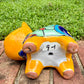 A hand-painted ceramic cat figurine in bright yellow with red, blue, and beige patterns, standing on a brick surface with a blurred green lawn and house in the background, bottom view.