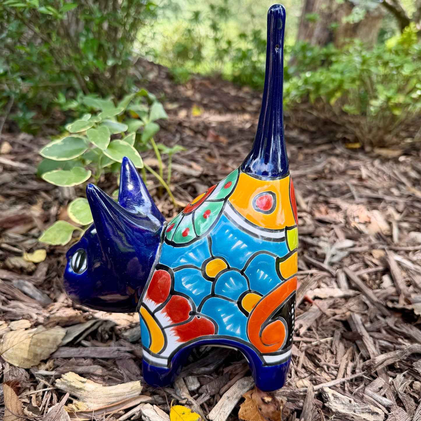 A colorful ceramic cat figurine with a glossy blue head and tail, yellow ears and paws, and a patterned body, standing outdoors on mulch with green plants in the background, right side view of cat.
