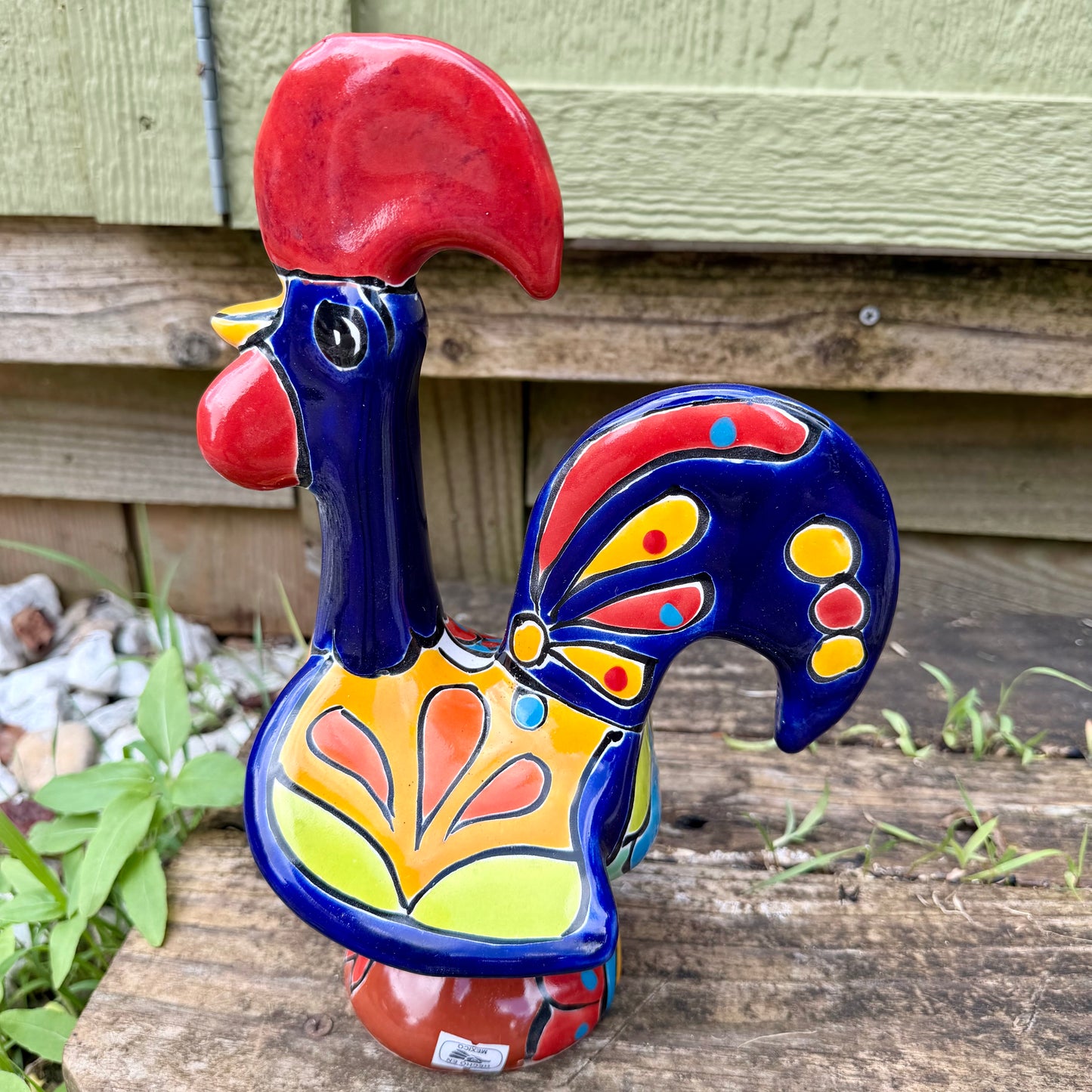 A colorful hand-painted ceramic rooster figurine standing on a rustic wooden surface in front of a green wooden door, right side view.