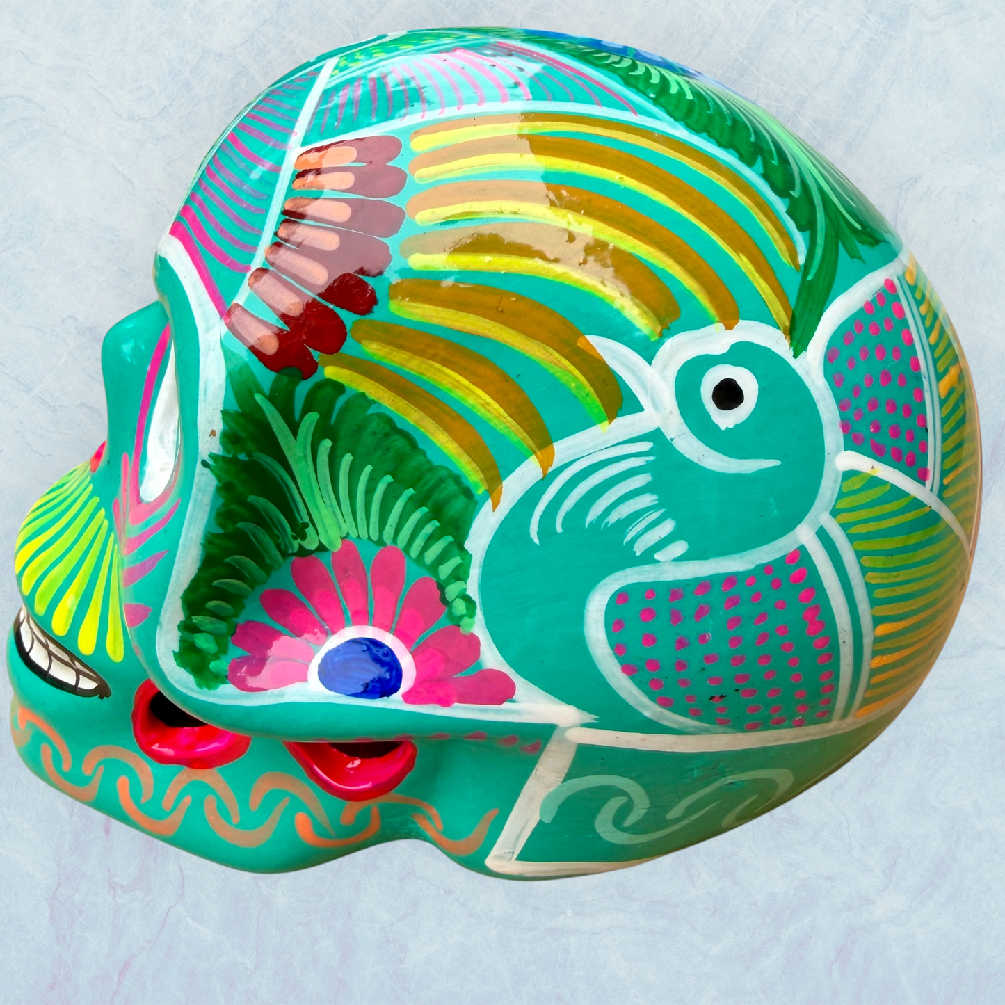 A decorative skull sculpture with intricate colorful sugar skull style patterns painted on a teal colored base, set against a light blue textured background, right side view.