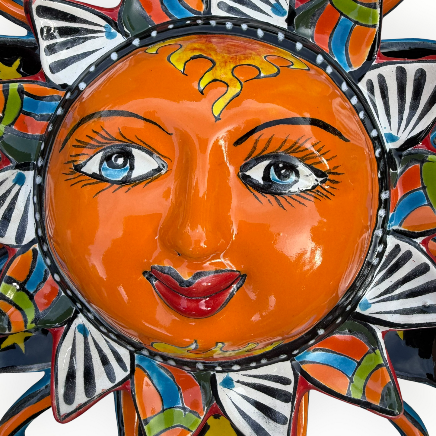A colorful hand-painted Talavera ceramic sun wall decor piece with a smiling face, orange center, planets, and multicolored rays on a plain white background face close up.