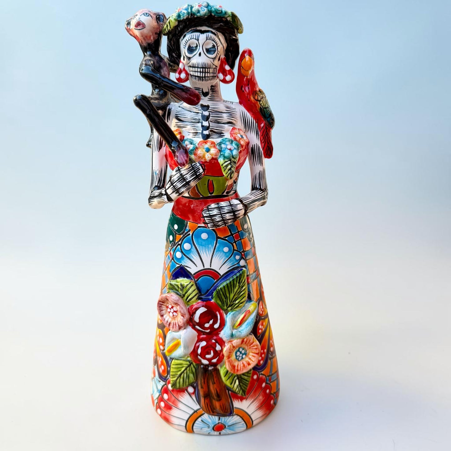 A Talavera Day of the Dead (Día de los Muertos) Frida figurine  with a monkey on her shoulder and a red parrot on her other shoulder, set against a light blue gradient background.