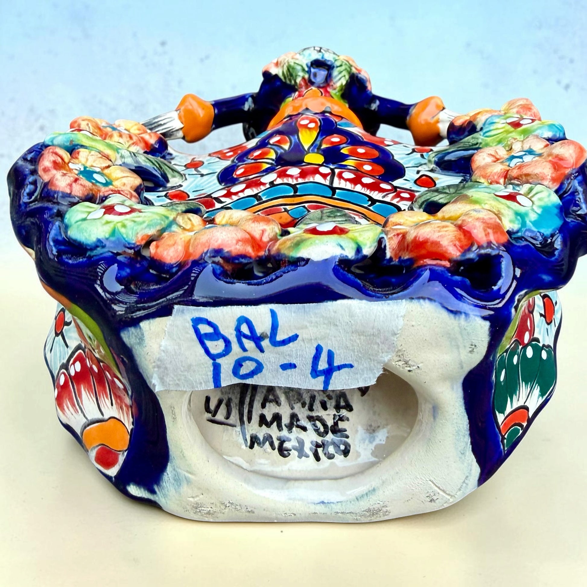 A colorful talavera Day of the Dead (Día de los Muertos) dancing skeleton ballerina figurine. The figurine features black hair in braids wearing a vibrant multicolor dress with floral patterns and orange accents, set against a light blue and beige gradient background, bottom view showing artist signature.
