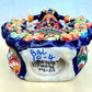 A colorful talavera Day of the Dead (Día de los Muertos) dancing skeleton ballerina figurine. The figurine features black hair in braids wearing a vibrant multicolor dress with floral patterns and orange accents, set against a light blue and beige gradient background, bottom view showing artist signature.
